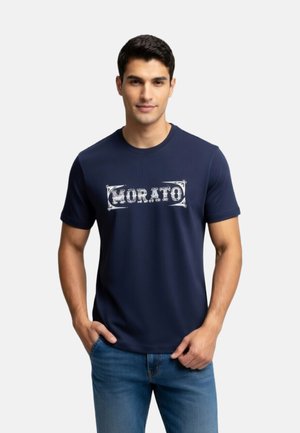 Hombre vistiendo una camiseta azul marino de manga corta con texto blanco "HORATO", combinada con vaqueros azules, de pie frente a un fondo liso y claro.