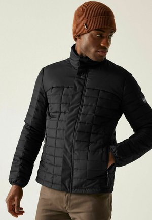 Mężczyzna w czarnej pikowanej kurtce i brązowej dzianinowej czapce, stojący z jedną ręką w kieszeni, patrzący w lewo.