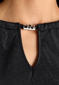 Svart topp med V-ringning och samlad halsringning. Har en silverfärgad detalj med utskärningar i mitten. Tyget har en subtil lyster.