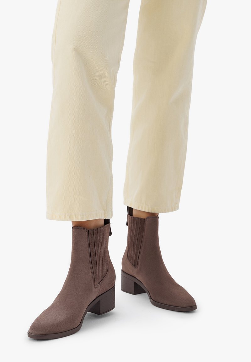 Piernas vistiendo pantalones beige claros y botas de ante marrón hasta el tobillo con tacones de bloque sobre un fondo blanco.