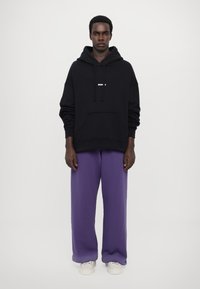 Črna kapuca z sprednjim žepom in belim napisom, v kombinaciji s prostornimi vijoličnimi trenirčkami in belimi supergami. Gladka tkanina, sproščen dizajn.