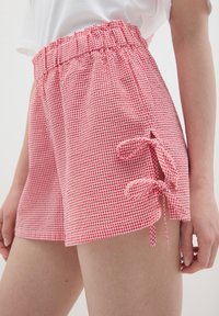 Shorts de cuadros rojos y blancos con cintura elástica, con aberturas laterales atadas con lazos de tela a juego. Material de algodón suave y ligero.