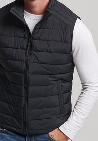 Schwarze Steppweste mit Reißverschluss, ausgestattet mit einem horizontalen Steppmuster, hohem Kragen und zwei seitlichen Taschen, getragen über einem weißen Langarmshirt.