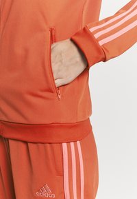 Main dans la poche zippée d'une veste de survêtement orange Adidas avec des bandes roses sur la manche et le pantalon, vue en gros plan.