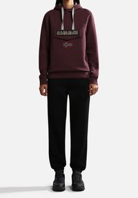 Sudadera burdeos con cordones, bolsillo delantero y parche del logotipo con la bandera noruega, combinada con pantalones negros y zapatillas negras.