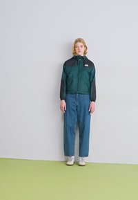 Tume teal ja must windbreaker jope, millel on kapuuts ja tõmblukuga esiosa, kombineeritud lõdva sinise teksapükste ja valgete kingadega.