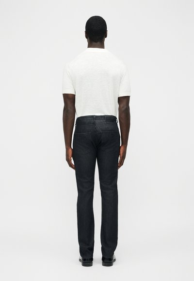 Άνδρας φαίνεται από πίσω φορώντας λευκό πόλο με κοντό μανίκι, σκούρο τζιν στενής γραμμής και μαύρα παπούτσια, στέκεται μπροστά σε απλό φόντο.