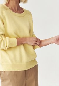 Żółty dzianinowy sweter z szerokim dekoltem i ściągaczami, połączony z beżowymi spodniami. Materiał wydaje się miękki o lekko teksturowanym wykończeniu.