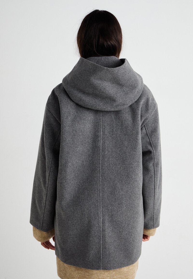 Szary wełniany płaszcz z dużym kapturem, opadającymi ramionami i prążkowanymi beżowymi mankietami na dole. Charakteryzuje się prostymi liniami i luźną sylwetką.