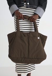 Borsa a spalla marrone trapuntata con doppio manico, dotata di una tasca frontale e un'etichetta con il logo. Ambientata su uno sfondo con una gonna a righe.