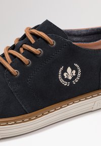 Sneaker in suede navy con lacci marroni, dettagli cuciti e un logo decorativo. La suola è beige con una finitura testurizzata.