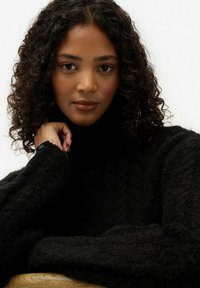 Czarny, strukturalny sweter z golfem o luźnym kroju. Posiada żebrowane szczegóły i lekko postrzępione mankiety. Model ma kręcone włosy i neutralny wyraz twarzy.
