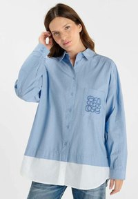 Lichtblauw gestreept oversized shirt met een witte zoom. Heeft een knoopsluiting aan de voorkant en een zak met een blauw grafisch ontwerp.