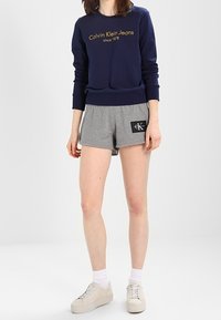Námorní modré mikina s zlatým textem "Calvin Klein Jeans", doplněná o světlé šedé kraťasy s černou nášivkou loga a bílými teniskami.