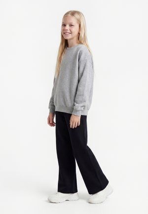 Ung pige med langt blond hår iført en grå sweatshirt, sorte bukser og hvide sneakers, smilende og stående i en afslappet positur.
