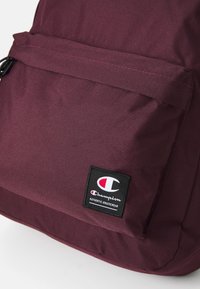Bordowy plecak z fakturowanej tkaniny. Posiada przednią kieszeń z czarną naszywką z logo Champion. Prosty design, bez dodatkowych ozdób.
