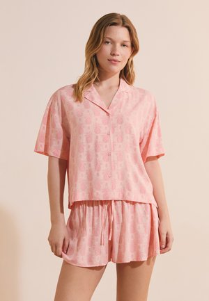 Donna che indossa una camicia rosa ampia a maniche corte con bottoni e pantaloncini abbinati con un delicato motivo di ananas, in piedi su uno sfondo beige.