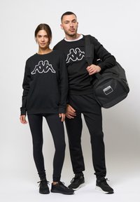 Zwarte sweatshirt met wit Kappa-logo, gecombineerd met zwarte leggings en sneakers. Het mannelijke model houdt een zwarte sporttas vast.