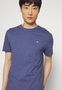 Ung mand iført en ensfarvet blå crewneck T-shirt med et lille broderet logo på venstre bryst, stående mod en ensfarvet baggrund.