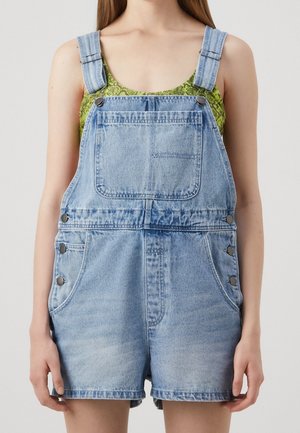 Jonge persoon draagt lichtblauwe denim korte tuinbroek met voorzak en groene top zonder mouwen met patroon.