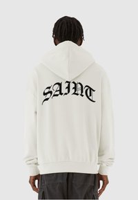 Weißes Hoodie mit großem schwarzem "SAINT"-Text auf dem Rücken. Verfügt über eine Kapuze mit Kordelzug sowie gerippte Bündchen und Saum. Weicher, strukturierter Stoff.