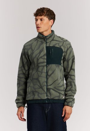 Joven viste chaqueta de forro polar verde con estampado, con bolsillo oscuro en el pecho y cremallera, de pie frente a un fondo claro y liso.