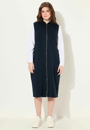 Robe sans manches bleu marine avec une fermeture éclair sur le devant, en tissu lisse, présentant un col haut et une silhouette midi, associée à des baskets blanches.