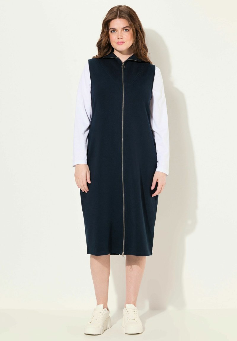 Abito senza maniche blu navy con zip frontale, realizzato in tessuto liscio, caratterizzato da un colletto alto e una silhouette fino al ginocchio, abbinato a sneakers bianche.