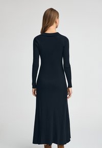 Svart ribbad stickad klänning med långa ärmar och åtsittande liv, som slutar vid ankelhöjd, med en subtil krage och mjuk textur.