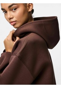 Felpa marrone con cappuccio ampio, texture morbida e dettagli di cucitura visibili. Il modello tiene il cappuccio vicino al viso.