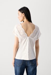 Chemisier blanc avec un dos en V profond, des détails en dentelle délicate avec des découpes circulaires, des manches courtes et une coupe décontractée, porté avec un jean bleu.