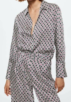 Femme portant un chemisier et un pantalon à motifs de losanges noirs, blancs et roses, avec une taille ceinturée et des manches longues.