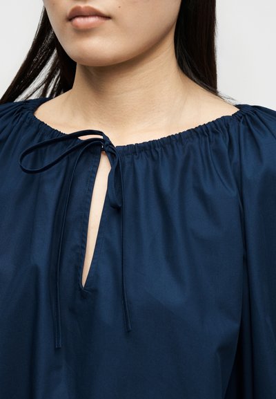 Γυναίκα που φοράει μπλε μπλούζα με στρογγυλή λαιμόκοψη που δένεται με φιόγκο μπροστά, αποκαλύπτοντας ένα μέρος από τον λαιμό και τα χείλη της.