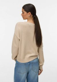 Femme aux longs cheveux noirs attachés en queue de cheval, portant un pull en maille beige et un jean bleu clair, vue de profil sur un fond blanc uni.