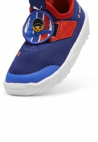 Navy sininen urheilujalkine, jossa on punaisia yksityiskohtia, teksturoitu kangas, pehmustettu kaulus ja pyöreä logo, jossa on sarjakuvahahmo. Valkoinen pohja.