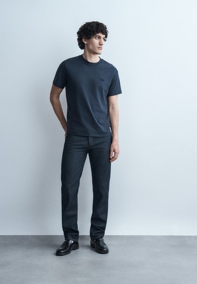 Jeune homme aux cheveux bouclés portant un t-shirt bleu marine, un jean foncé et des chaussures noires, debout devant un mur gris clair.