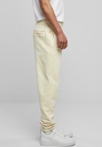 Light beige sweatpants with a tapered fit, elastic waistband, side pocket, and ribbed cuffs, paired with white sneakers.