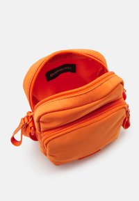 Oransje stoff crossover-veske med glidelås. Har to rom, myk tekstur og et merkeinnslag inni. Kompakt design.