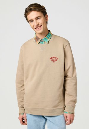 Beżowa bluza z wyszywaniem "WRANGLER JEANS 1947" w kolorze czerwonym, okrągły dekolt oraz ściągacze przy rękawach. Nosi na sobie wielokolorową koszulę w kratę.