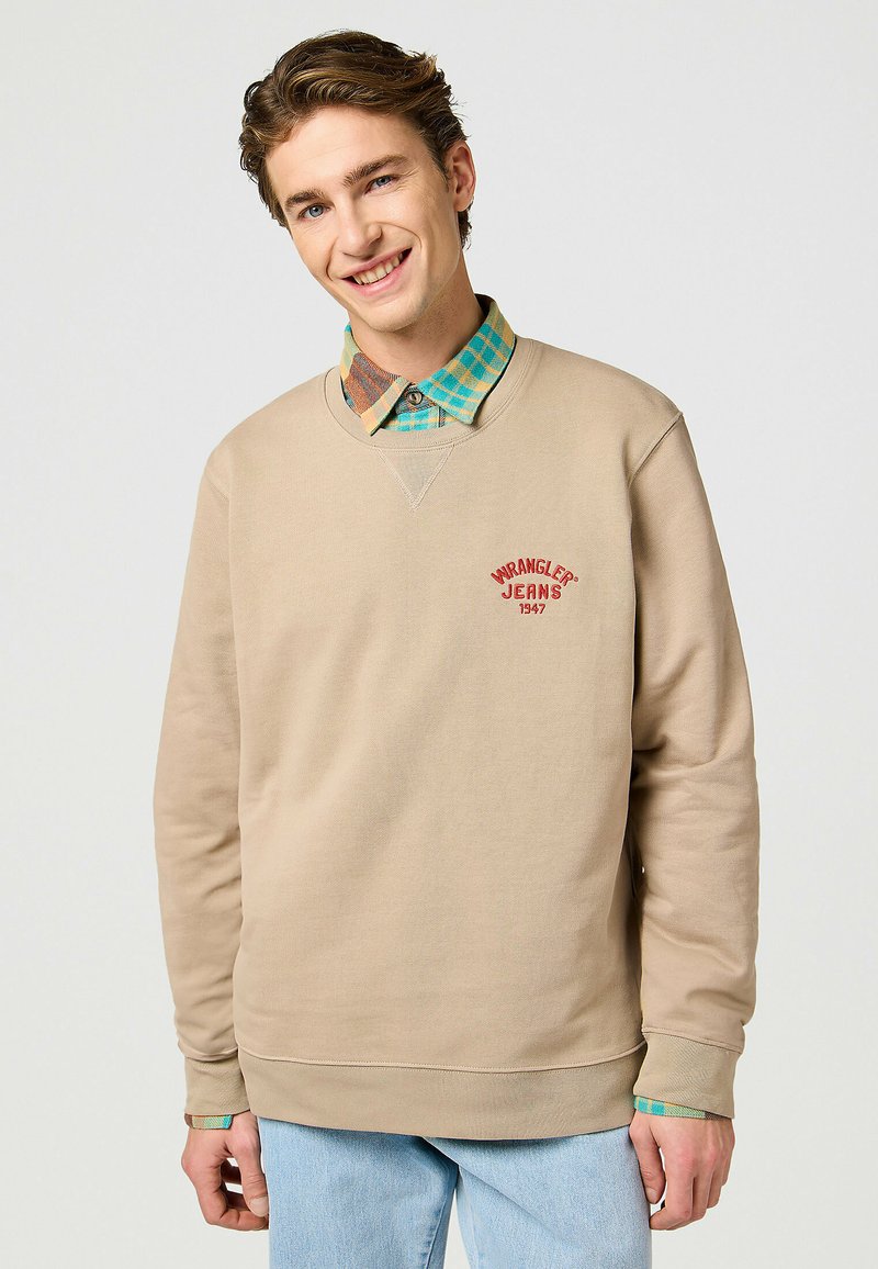 Felpa beige con ricamo rosso "WRANGLER JEANS 1947", scollatura rotonda e polsini a coste. Indossata sopra una camicia a quadri multicolore.