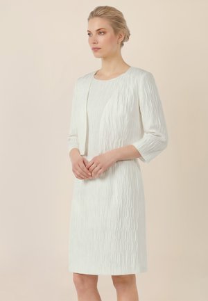 Frau mit blondem Haar zu einem Hochsteckfrisur gebunden, trägt ein strukturiertes weißes knielanges Kleid und eine passende kurze Jacke, steht mit gefalteten Händen.