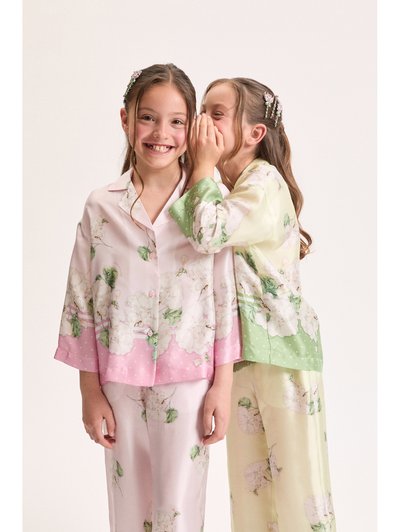 Δύο κορίτσια με κλιπ μαλλιών σε σχήμα λουλουδιού φορούν πυτζάμες με λουλούδια· η μία ψιθυρίζει στην άλλη, που χαμογελά στην κάμερα μπροστά σε απλό φόντο.