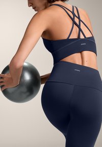 Zestaw do ćwiczeń w kolorze granatowym, składający się z sportowego biustonosza z paskami oraz legginsów z wysokim stanem, wykonany z gładkiego, elastycznego materiału. Trzymająca metaliczno-szary ball.