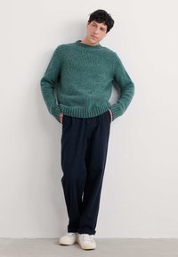 Tirkizno pleteni pulover s rebrastim okruglim izrezom, uparen s ležernim tamnoplavim hlačama i bijelim tenisicama, na neutralnoj pozadini.