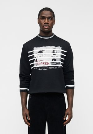 Homme portant un sweat-shirt noir avec des rayures blanches et un graphisme noir et blanc ressemblant à une photo d'identité judiciaire avec le texte rouge « BUSTED ».
