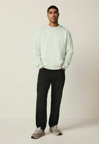 Joven de pie con las manos en los bolsillos, vistiendo una sudadera de color menta claro, jeans negros y zapatillas beige contra un fondo liso.