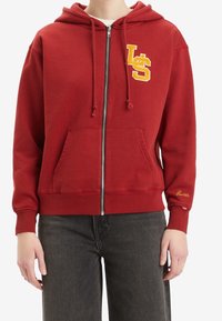 Sudadera azul con cremallera, hecha de algodón, con capucha, bolsillos frontales y logo bordado. Presenta letras amarillas en contraste y detalles de costura sutil.