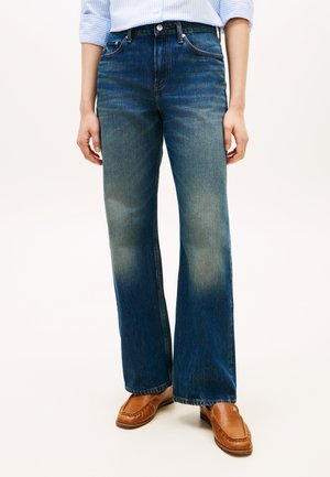 Person iført blå jeans med lige ben, lyse falmede områder på lår og knæ, kombineret med lysebrune læderloafers.