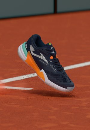 Dunkelblauer und oranger Tennisschuh schwebt in der Luft über einem roten Sandplatz, wirbelt Tonpartikel in der Nähe der weißen Begrenzungslinie auf.