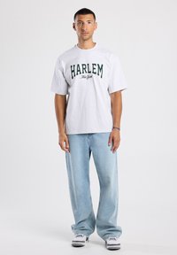 T-shirt en coton gris avec "HARLEM" et "New York" imprimés en vert. Associé à un jean en denim bleu clair et décontracté et des baskets noir et blanc.